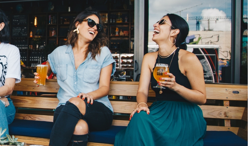 Two women holding drinks laugh sitting outside a restaurant 